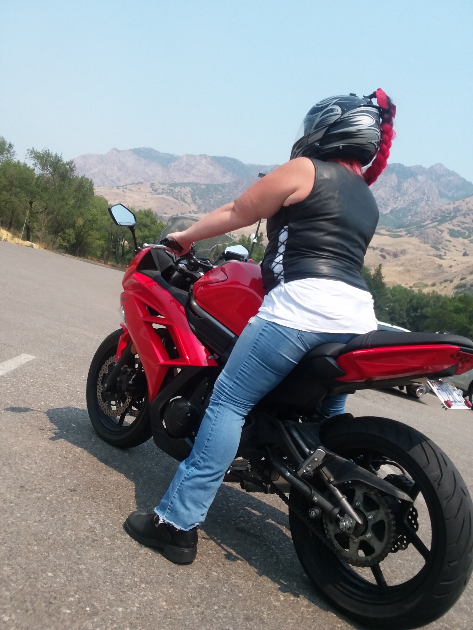 A woman sits astride a red Kawasaki Ninja 650, quartering away from the foreground so that her back, left arm, left hip and left leg are visible. She wears blue jeans, black boots, a white tank top and a leather vest. The white tank top shows through laces in the side of the vest. She also wears a black and gray full-face helmet, the crown adorned with a long faux ponytail of braided red fiber. 